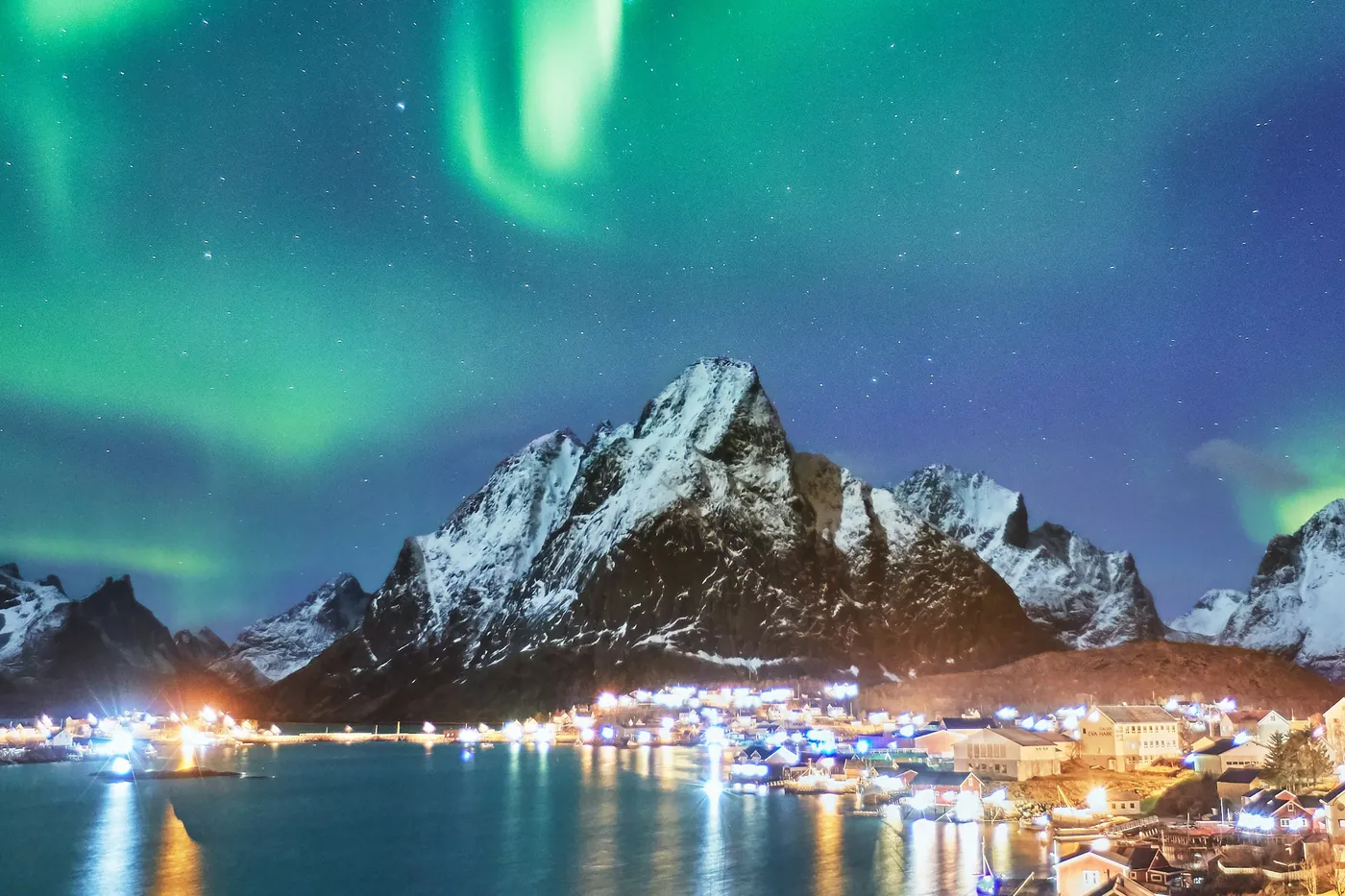 Grüne Nordlichter über einem schneebedeckten Gipfel und dem beleuchteten Dorf Reine, das sich in der Bucht spiegelt