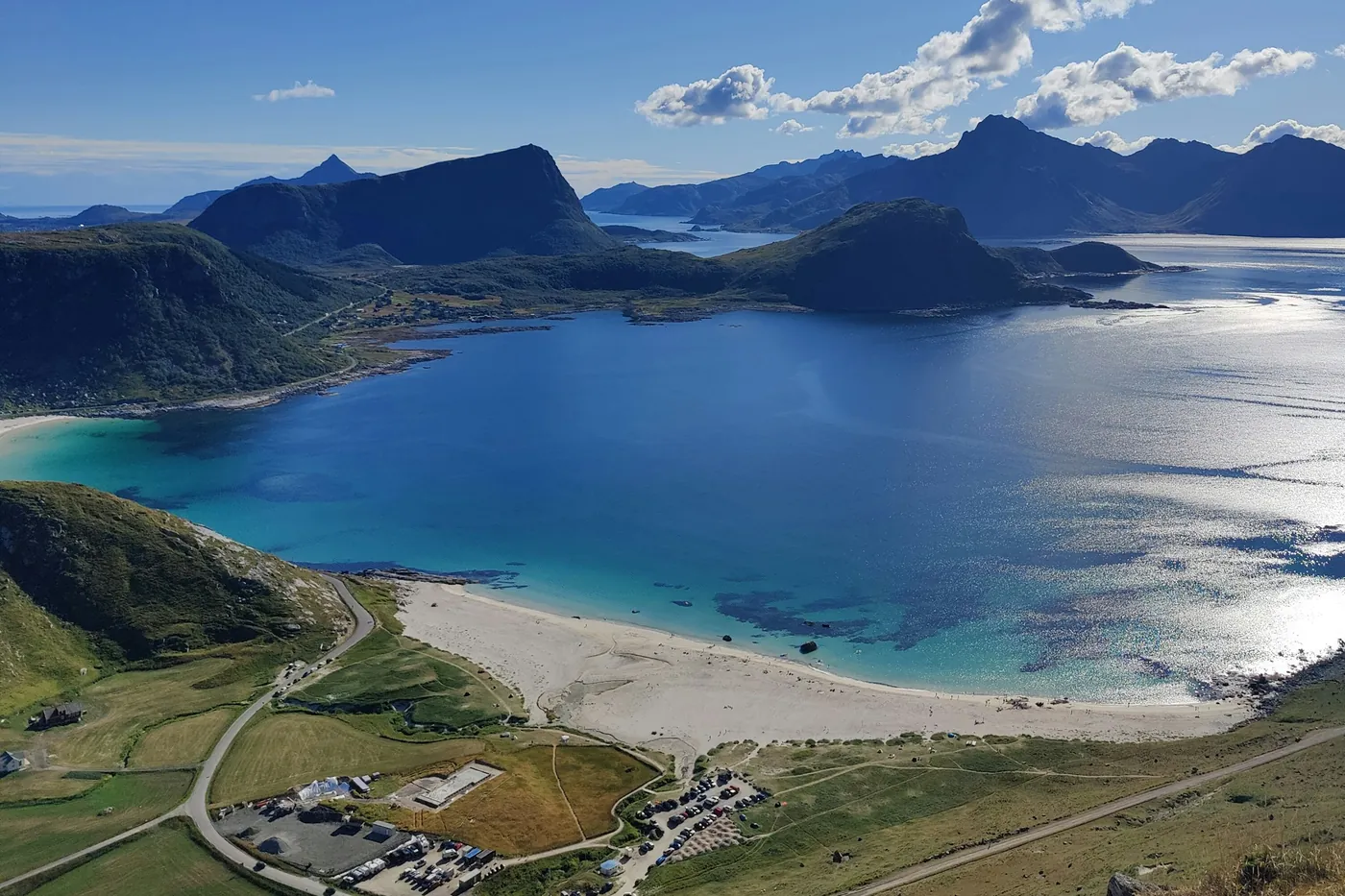 Vue aérienne d’une plage de sable blanc et d’une baie turquoise avec des montagnes, une route et des champs au premier plan