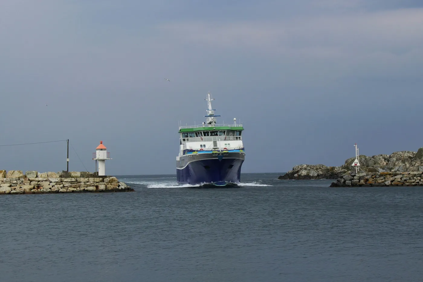 Ferry qui arrive entre deux brise-lames avec un petit phare, sous un ciel gris