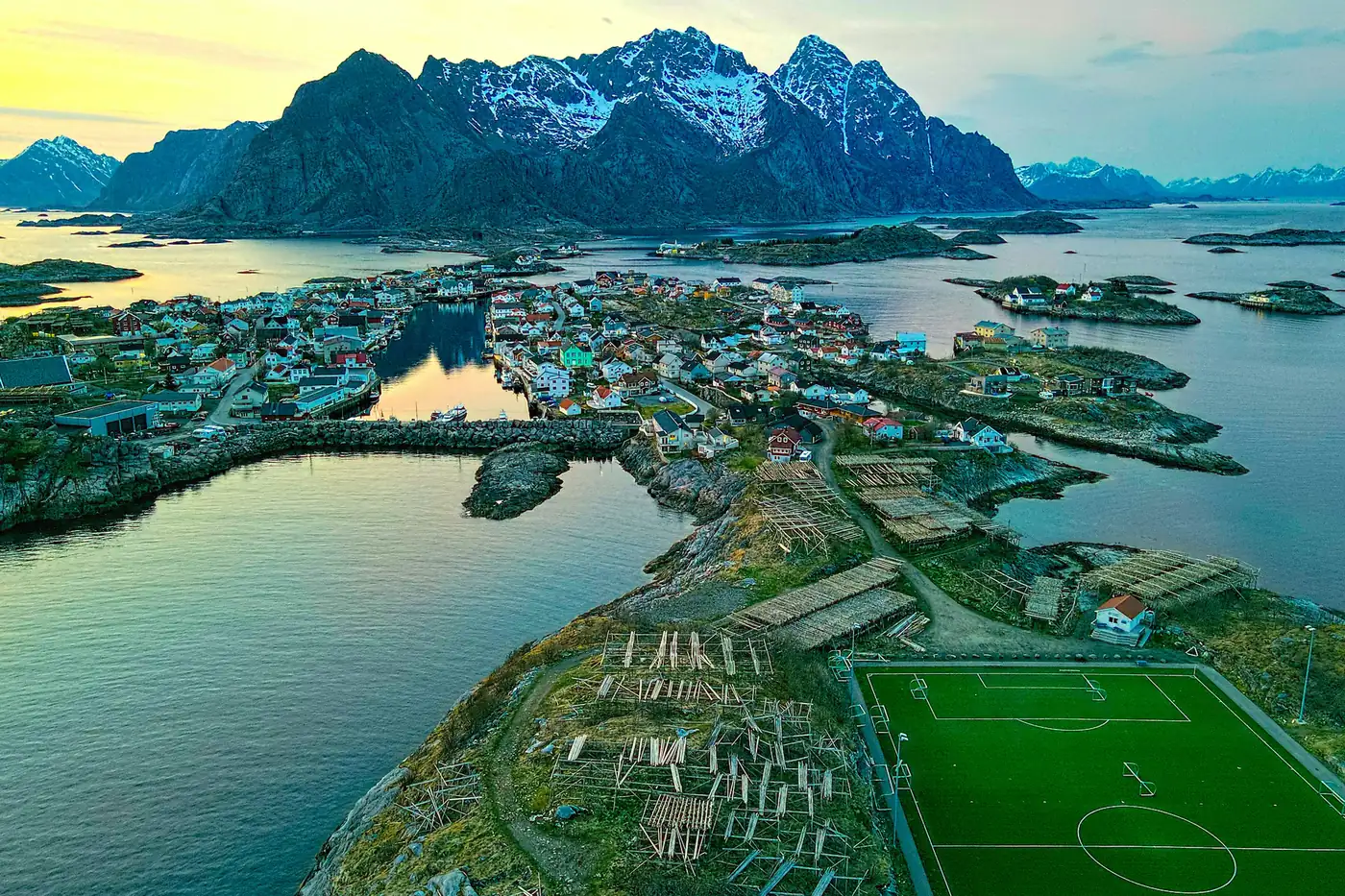 Luftbild von Henningsvær mit Fußballplatz, Fischtrockengestellen und Häusern, dahinter Berge