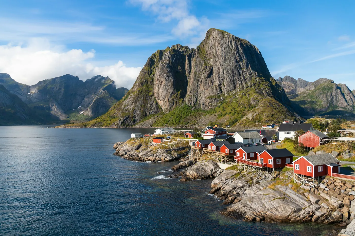 Cabañas rorbu rojas y blancas en una orilla rocosa de Hamnøy, bajo un pico de granito empinado y con el agua azul en calma