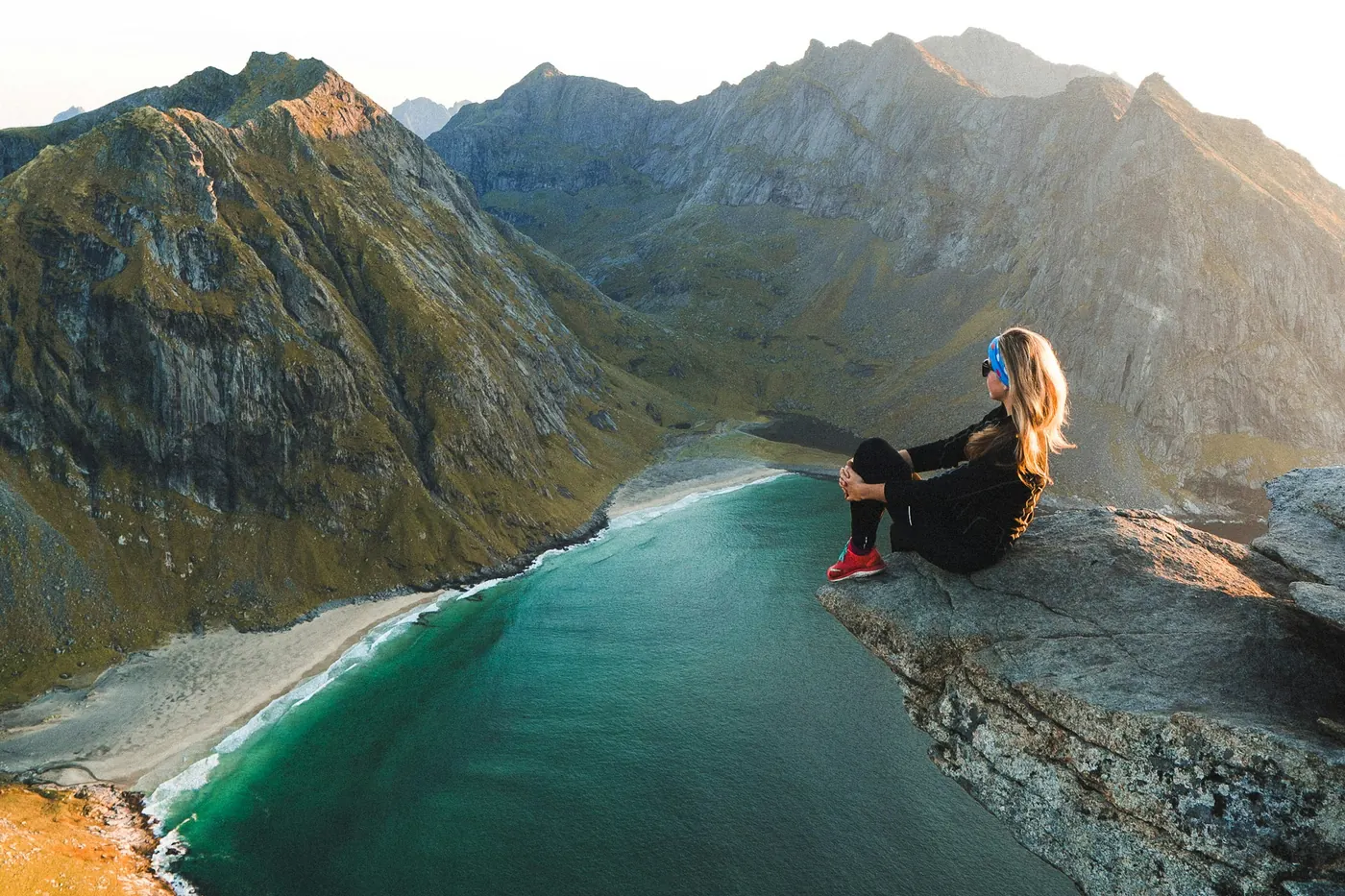 Wanderer sitzt auf einem Felsvorsprung über einer türkisfarbenen Bucht und einem Sandstrand zwischen Bergen