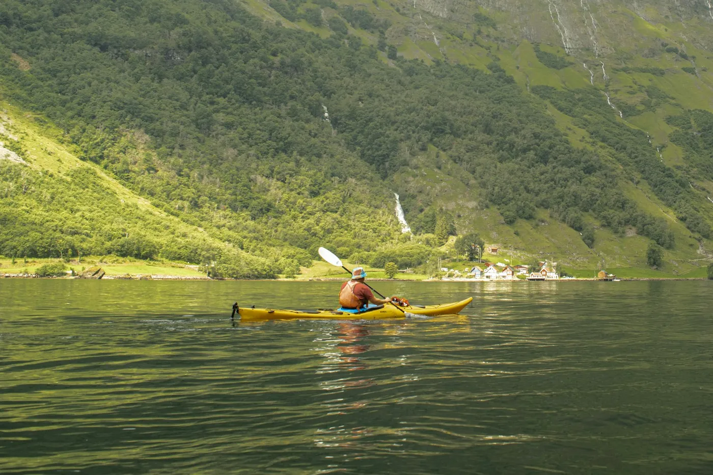 Kayaker in a yellow kayak on green fjord water below steep forested slopes with waterfalls