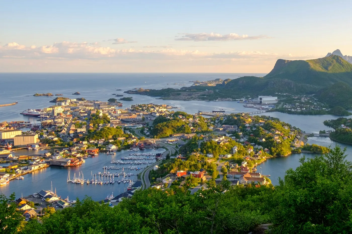 Die Stadt Svolvær und ihr Hafen auf kleinen Inseln, von einem Hang im Abendlicht gesehen, dahinter Berge