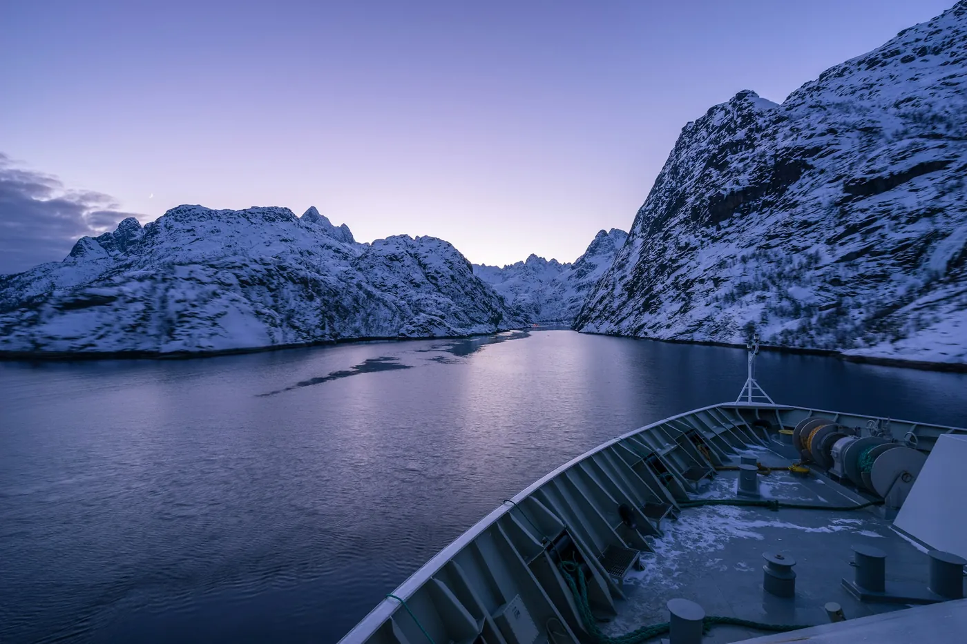 Bug eines Schiffs bei der Einfahrt in einen schmalen Fjord zwischen steilen, verschneiten Bergwänden in der Dämmerung