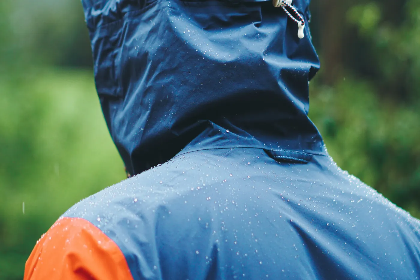 Person in blau-oranger Regenjacke mit Kapuze von hinten, Regentropfen auf dem Stoff